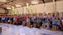 Messehalle mit Menschen auf Papphockern, die den Arm zur Abstimmung gehoben haben. Resolutionsabstimmung beim Kirchentag in Dortmund 2019. (Foto: Kirchentag/ Benedikt Bahl)